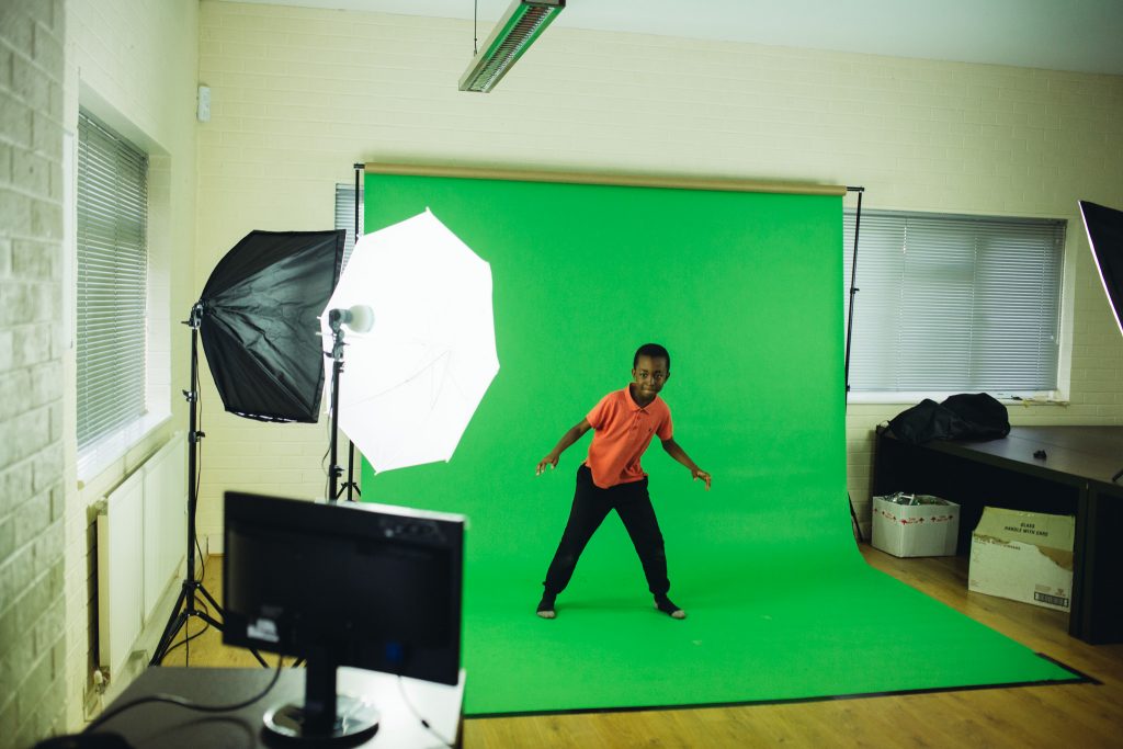 Image depicts a young Black boy with fliming equipment posing in front of a greenscreen
