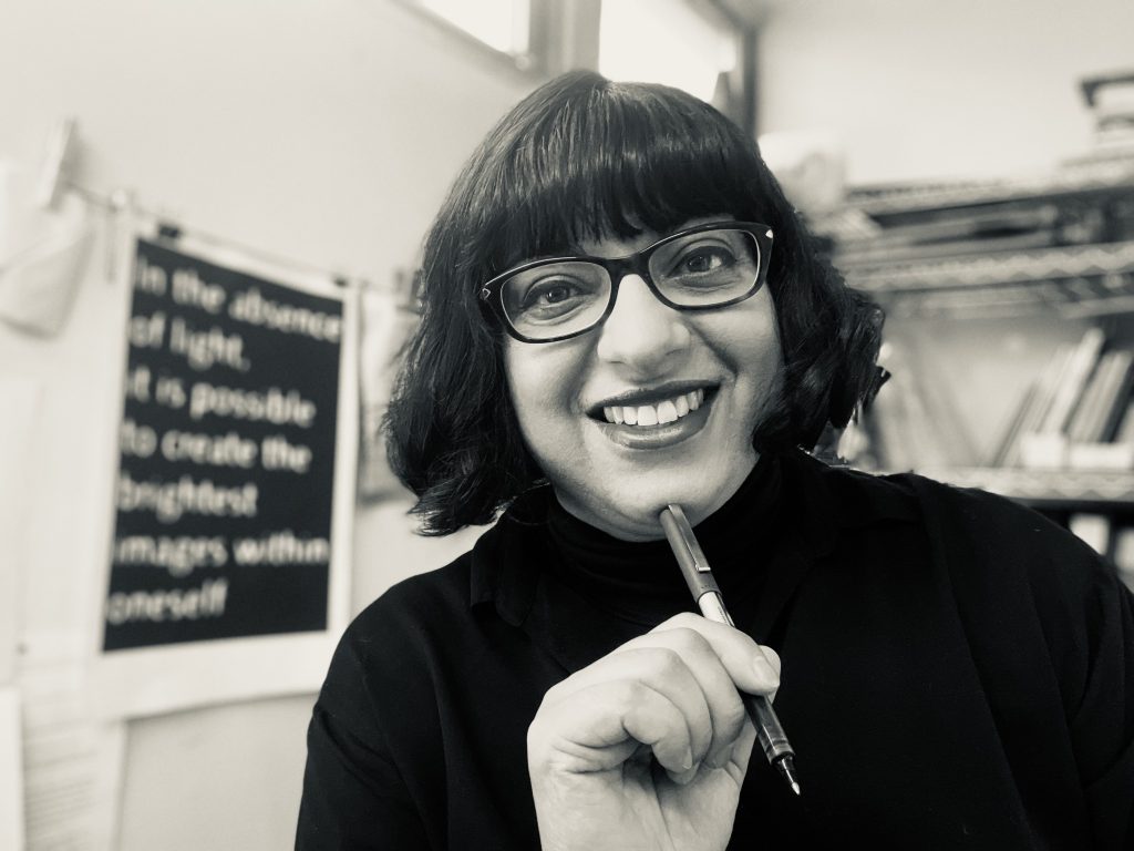 A black and white portrait of an indian woman with dark bobbed hair and glasses, smiling and holding a pen.