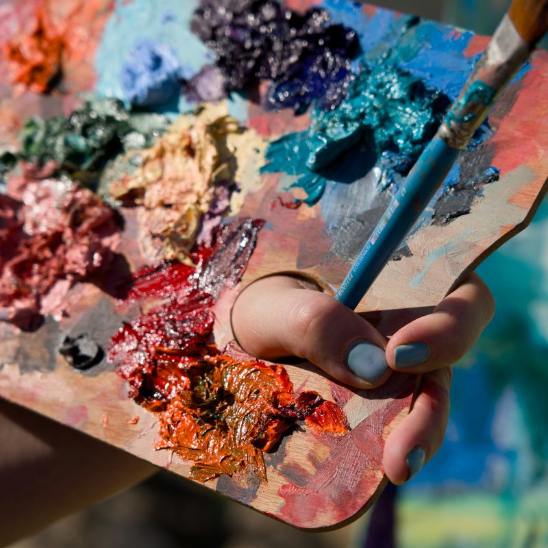 A hand holding a paint palette filled with paint with a paintbrush.