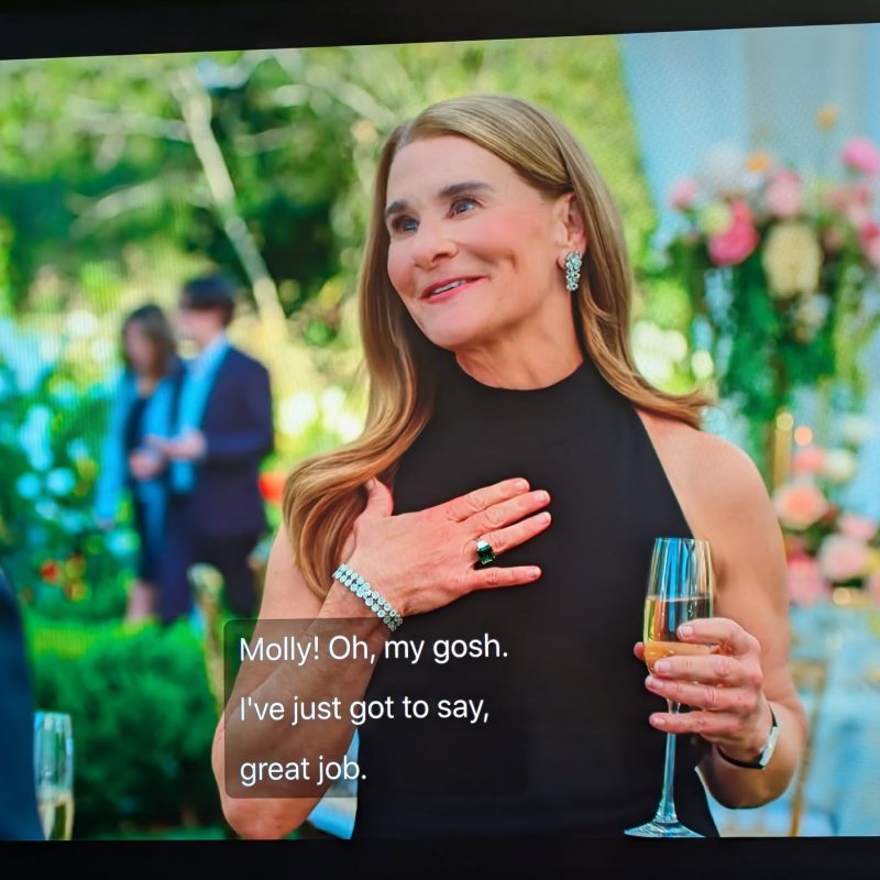 Melinda Gates appears on screen in a dress at a wedding holding a glass of champagne smiling